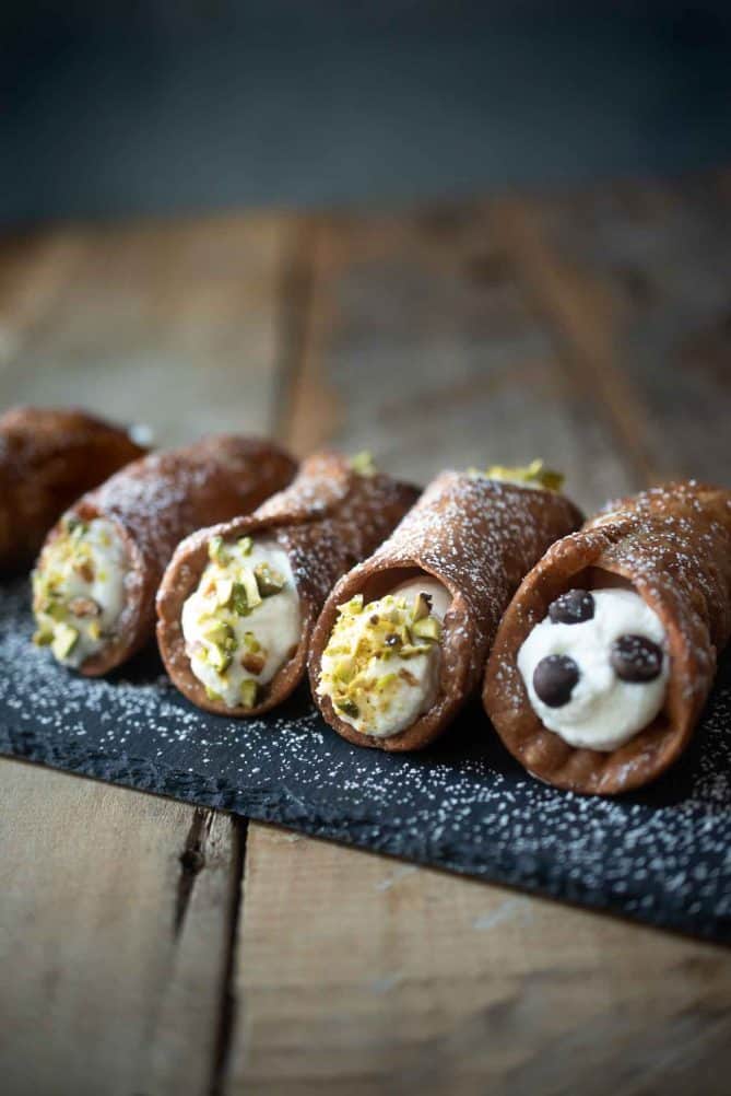 Cannoli lined up on a slate board, with chopped pistachios and chocolate chips