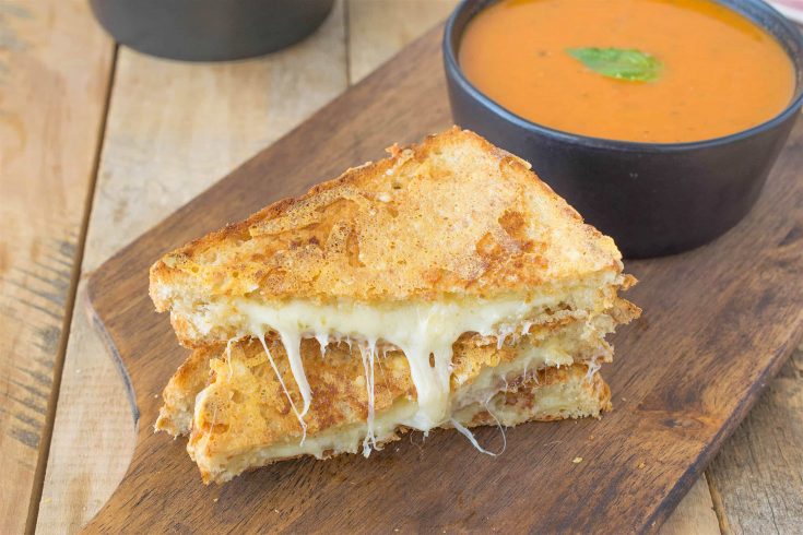 A triple grilled cheese sliced in half showing the melted cheese inside on a serving board with tomato basil soup