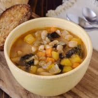 An off white bowl filled with Tuscan bean soup with bread, Parmesan and spoons