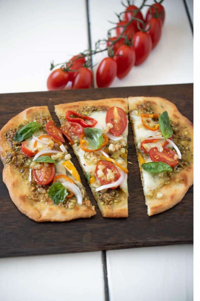 Vegetale pesto flatbread pizza cut into 3 slices on a board with cherry tomatoes on the vine