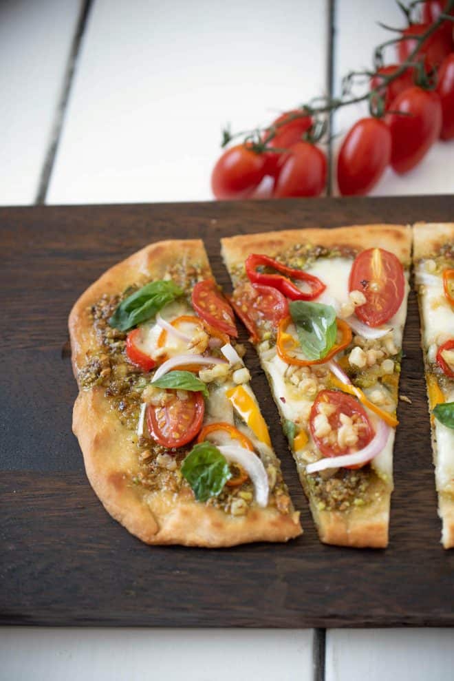 A closeup of a slice of vegetable pesto flatbread pizza showing the cherry tomatoes, red onion, corn, peppers, pesto and fresh basil
