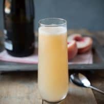 Prosecco and peach puree mixed in a stemless tall Champagne glass with a bottle of Prosecco and peach halves