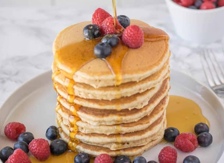 A large stack of pancakes topped with fruit pouring over syrup