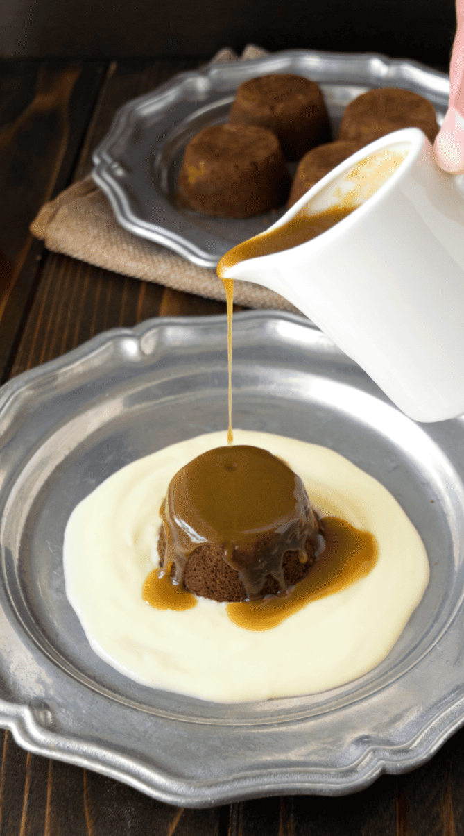 Dark brown toffee sauce on top of a round pudding with custard in the center of a plate